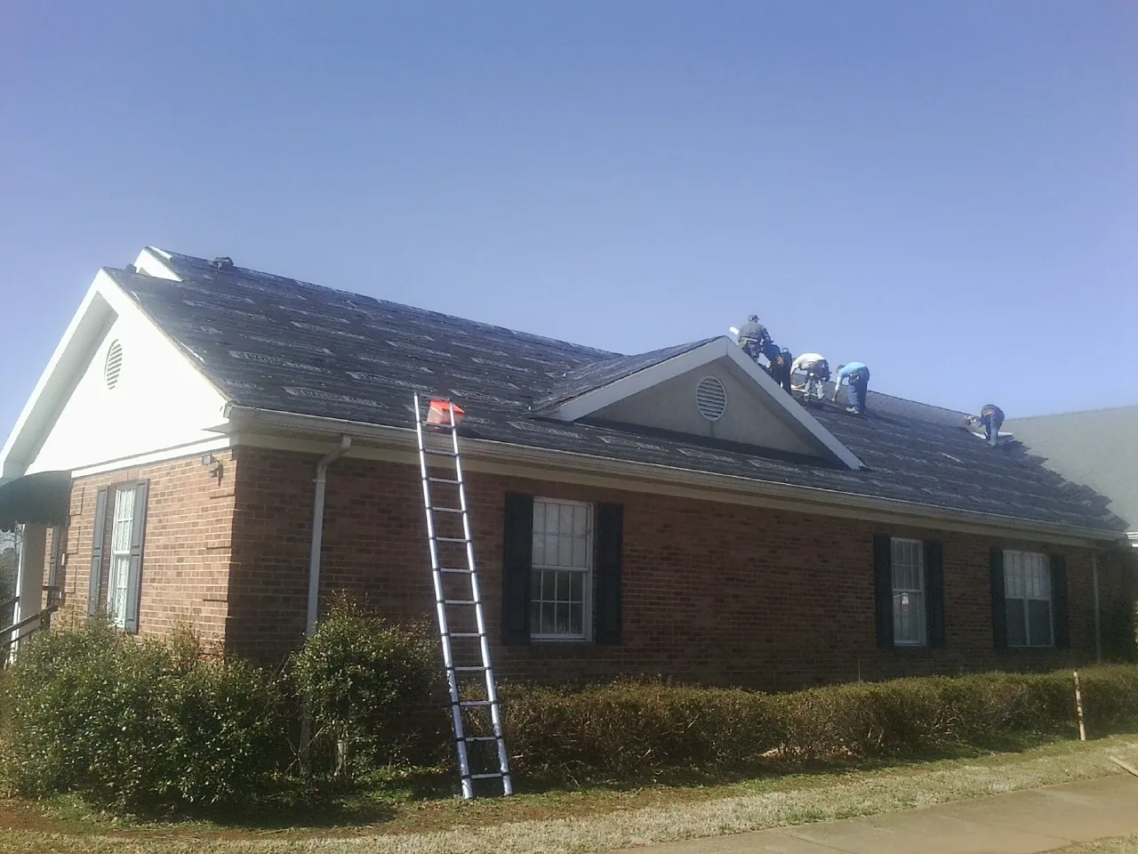 Crew tearing shingles off a brick ranch roof with a ladder set — Metal Roof Repair work by our crew in Barstow