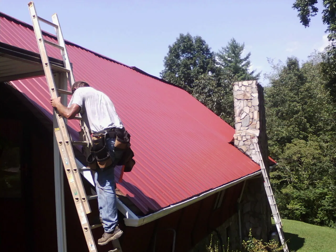 Roofer on a ladder beside a red metal roof and stone chimney — Soffit & Fascia Repair by Roofing Brothers in Barstow